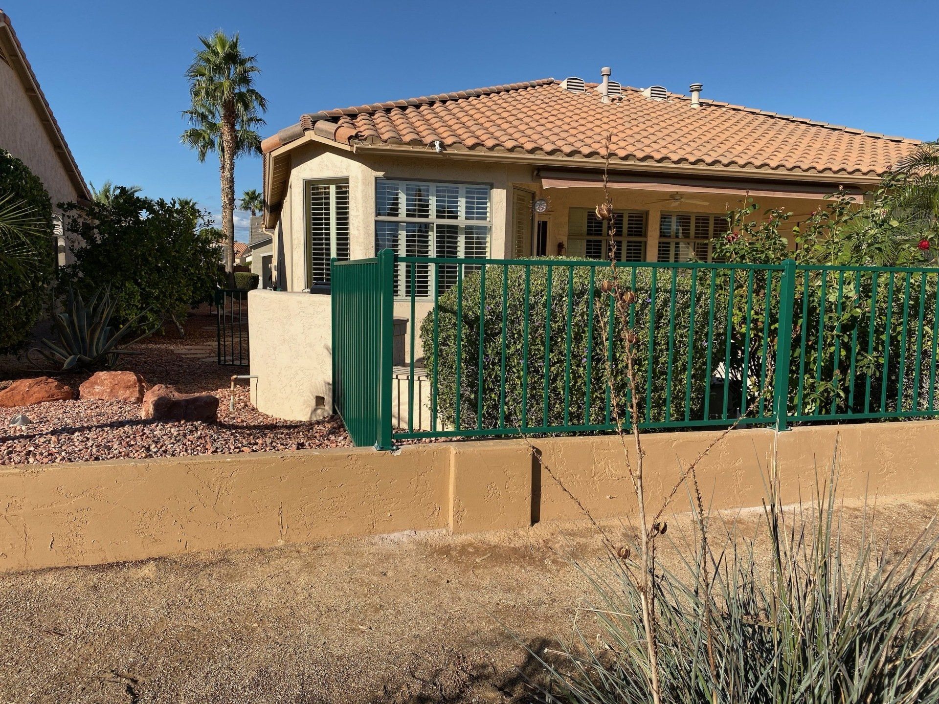 Tan stucco house with a green fence, and a tile roof on a sunny day.