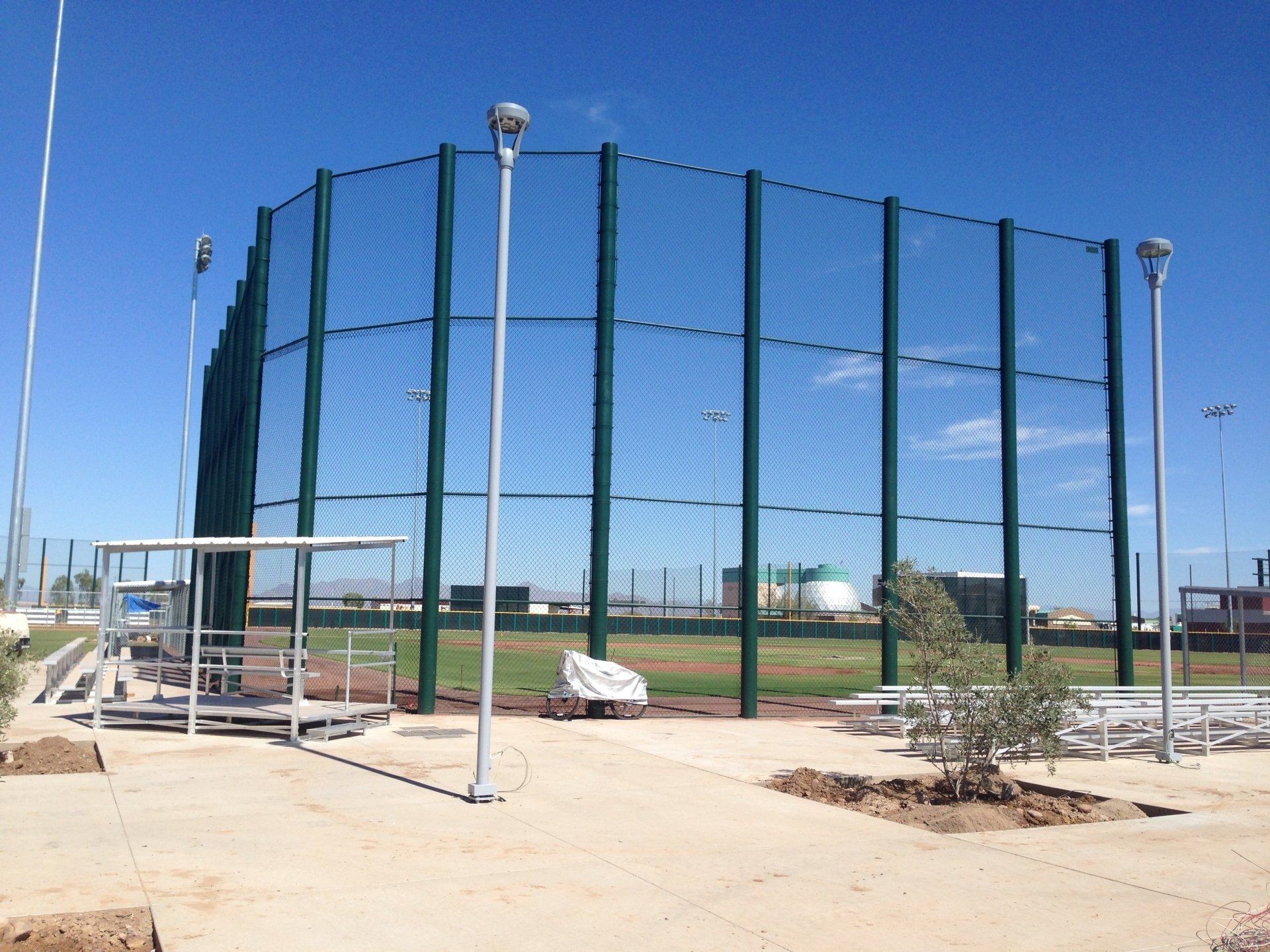 Baseball field with green netting, metal poles, and lights, under a clear blue sky.