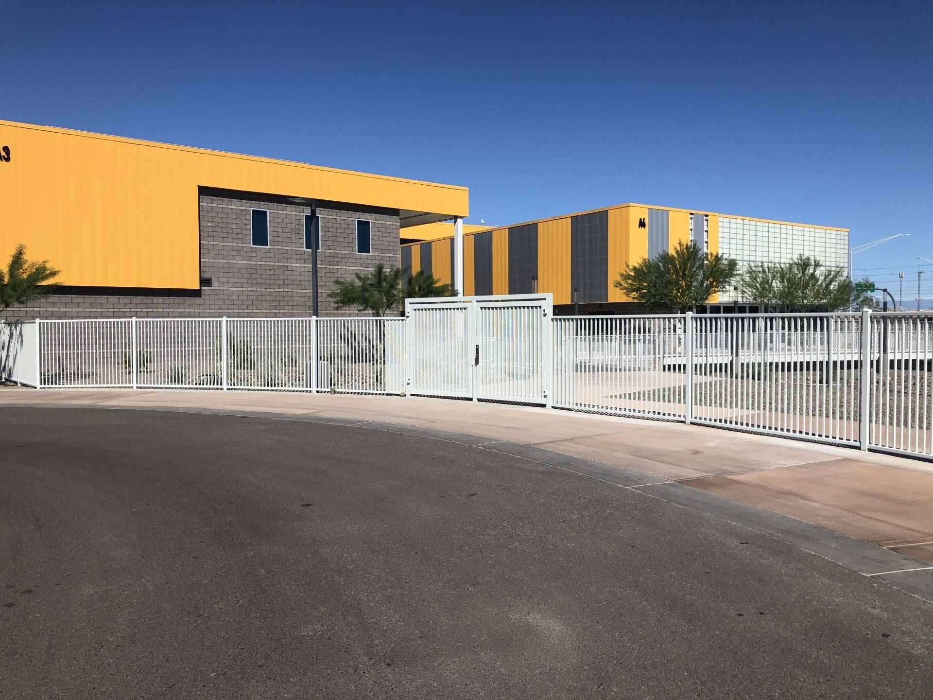 White gated entrance to a modern building with yellow and gray walls, under a clear blue sky.