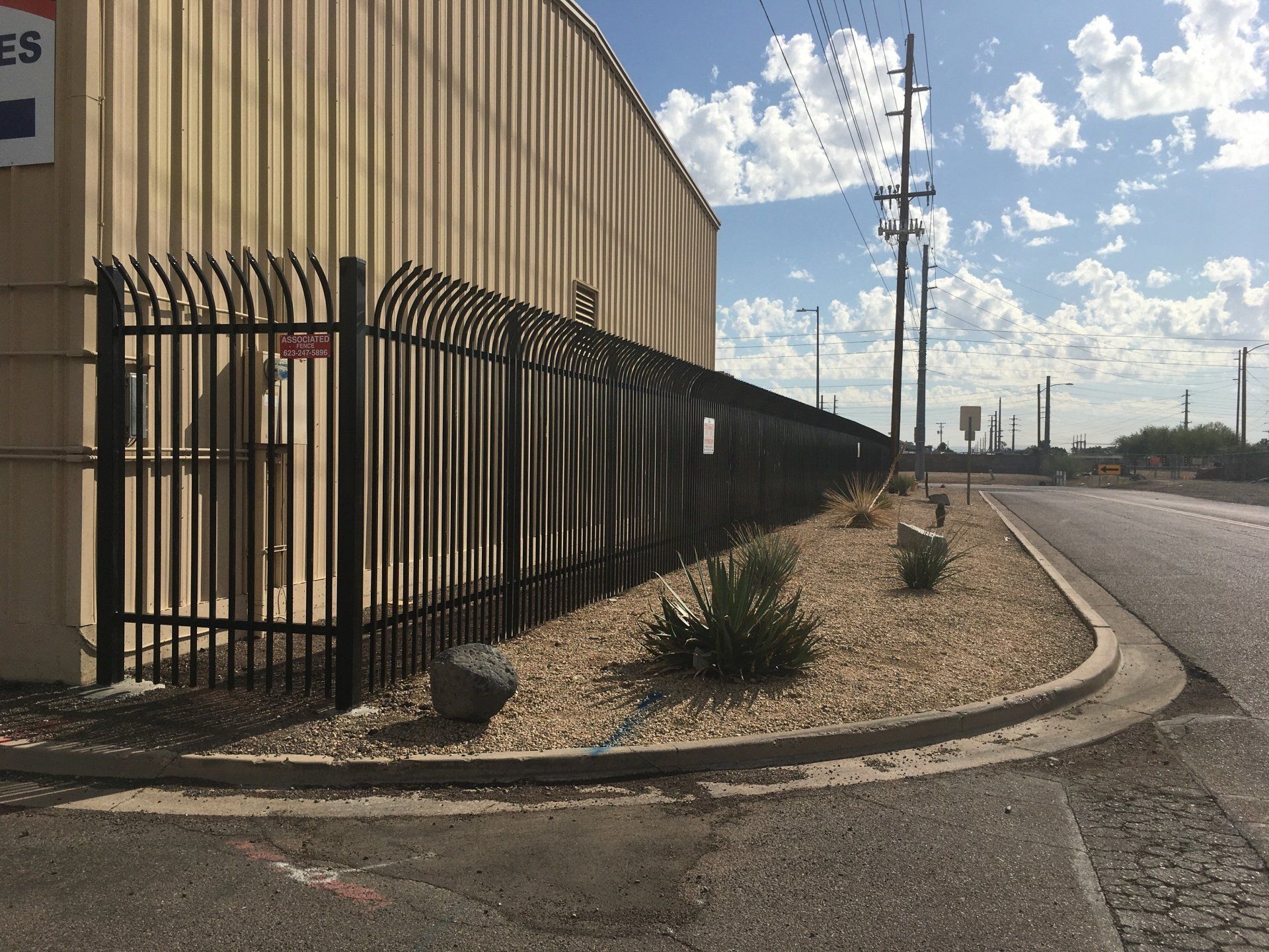 Black metal security fence alongside a beige building and a road with a blue sky.