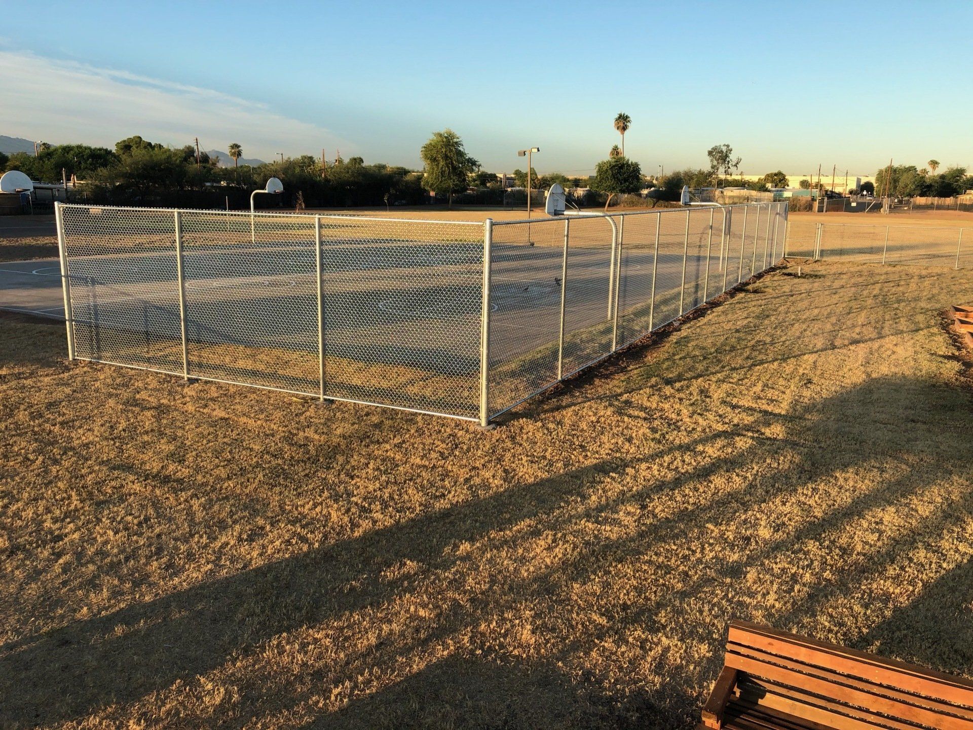 A basketball court enclosed by a metal fence on a grassy field in the sunlight.