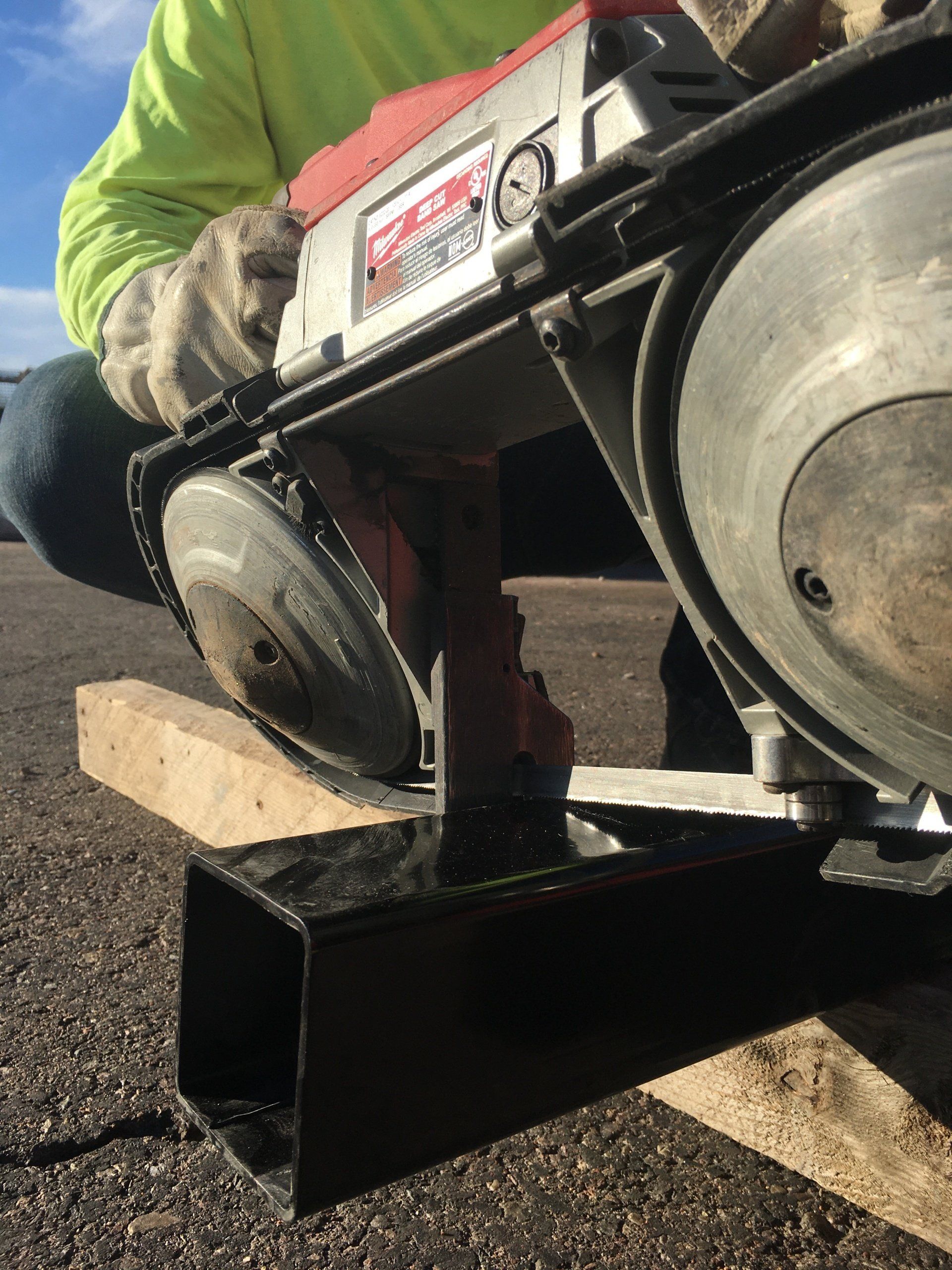 Person using a portable bandsaw to cut a black metal tube, outdoors.