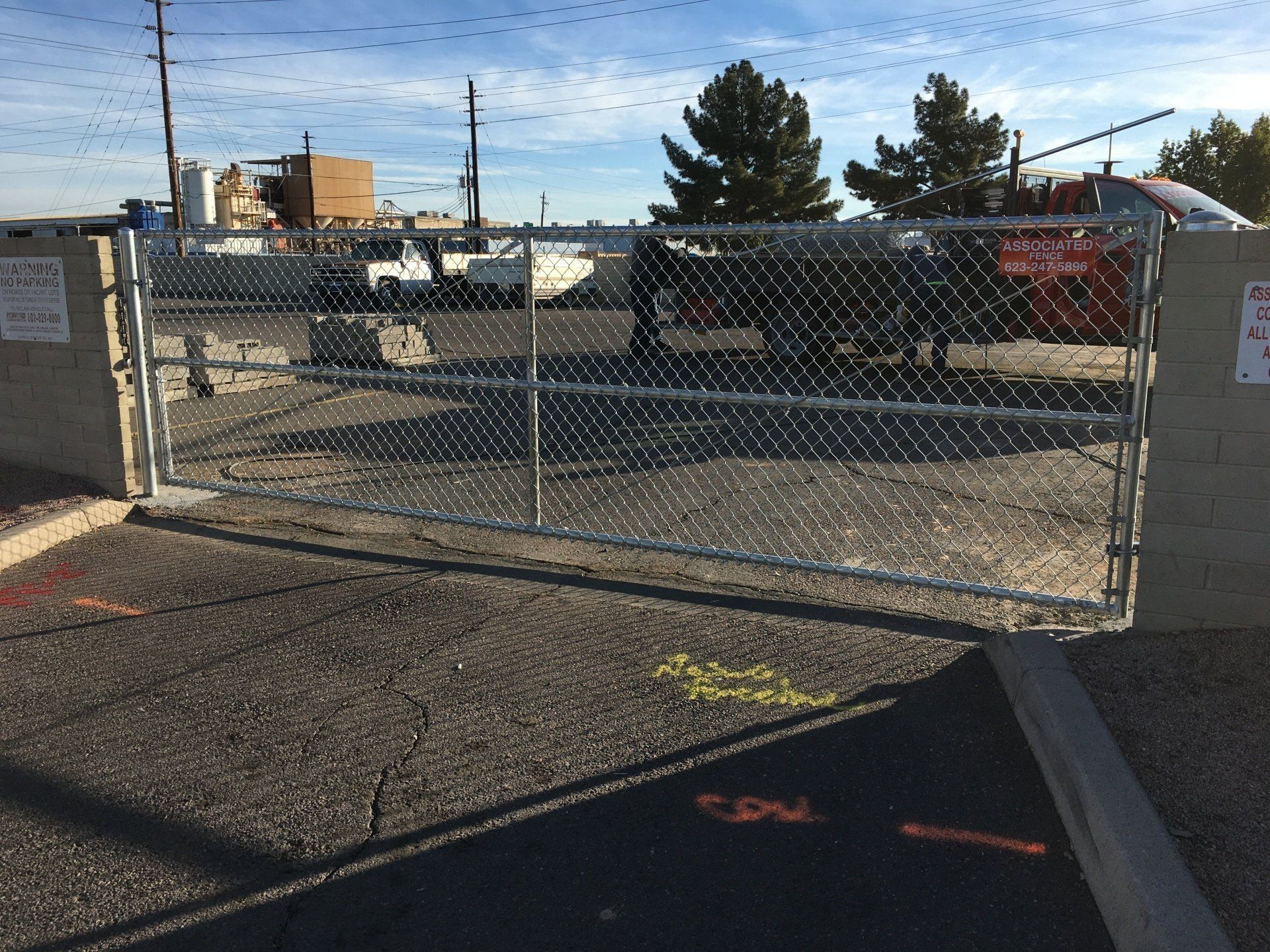 Chain-link gate in asphalt, leading to an industrial area. Concrete blocks flank it, with signs. Bright sunlight.