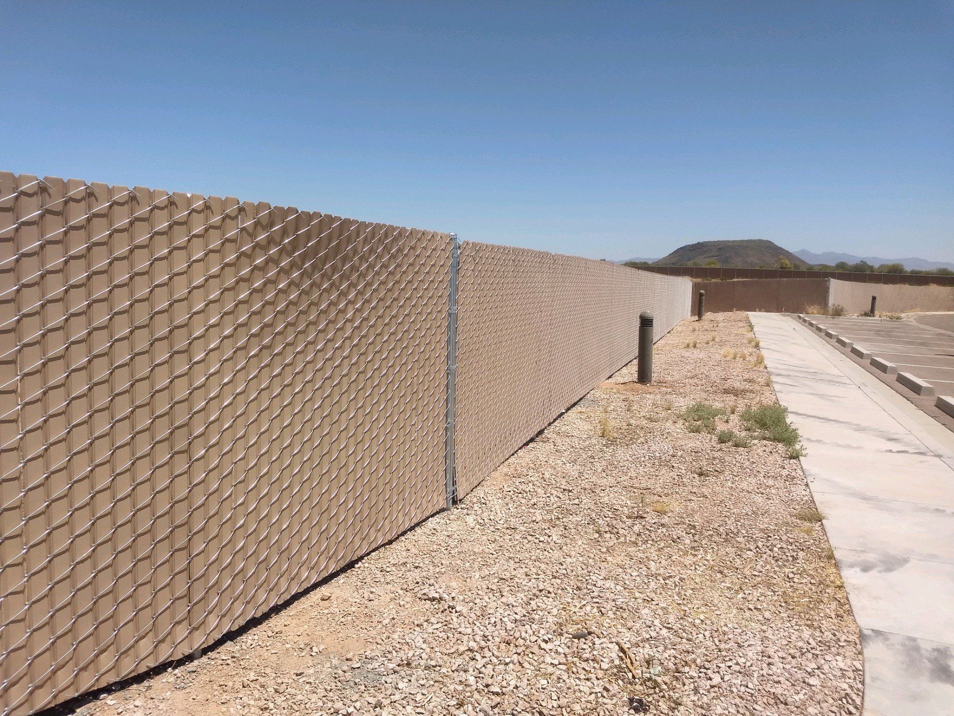 Tan chain-link fence on gravel next to a concrete path; blue sky.