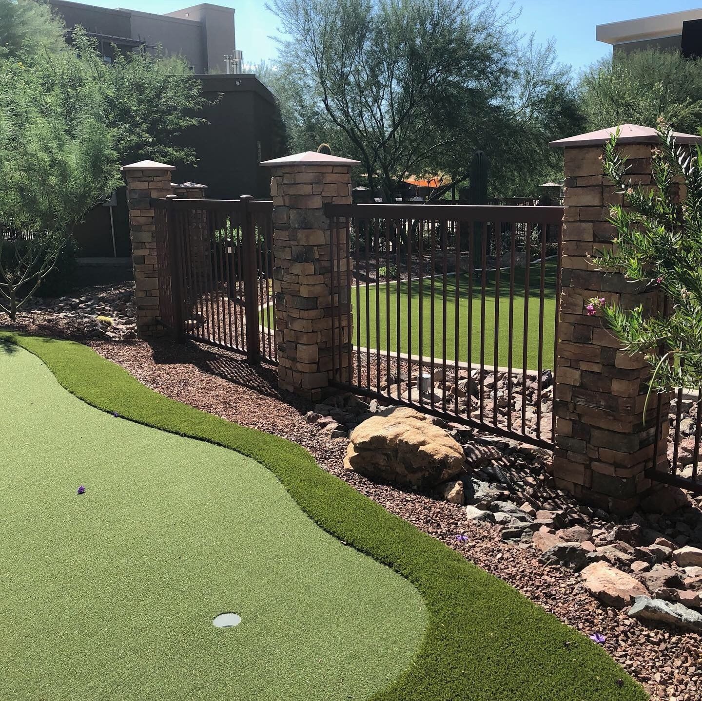 A backyard putting green with a stone and metal fence, and a brown rock border.