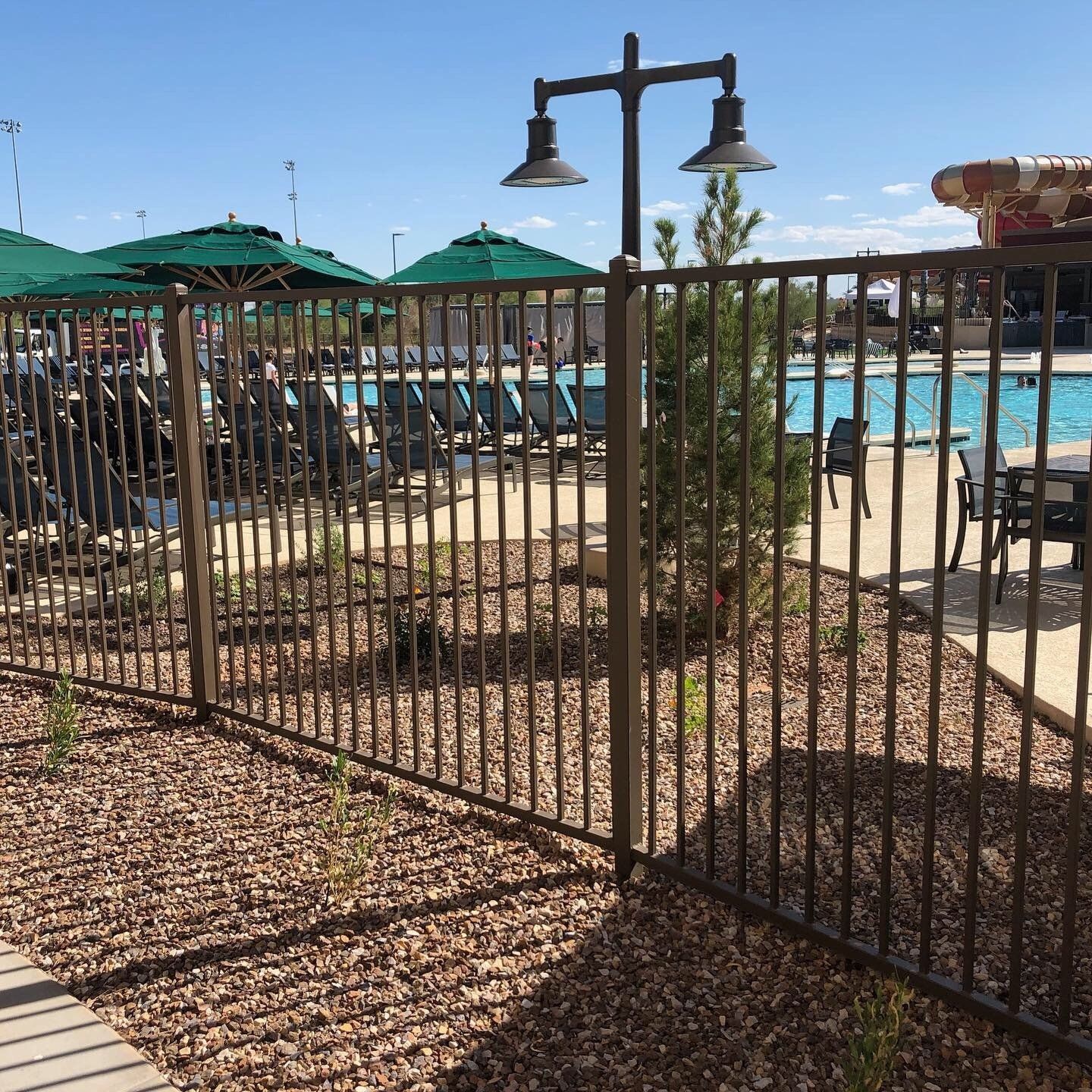 Brown metal fence surrounding a pool area with lounge chairs and umbrellas on a sunny day.