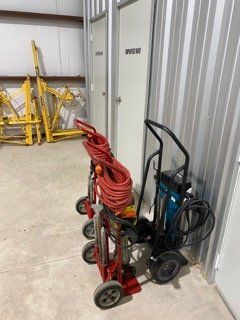 Red and black hand trucks with coiled hoses stored against a corrugated metal wall in a warehouse.