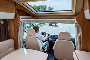 Interior view of a motorhome; person driving on road. Light interior, open sunroof.
