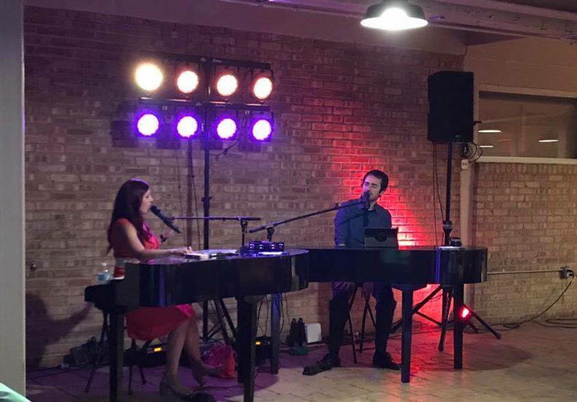 A man and a woman are sitting at a piano and singing into microphones.