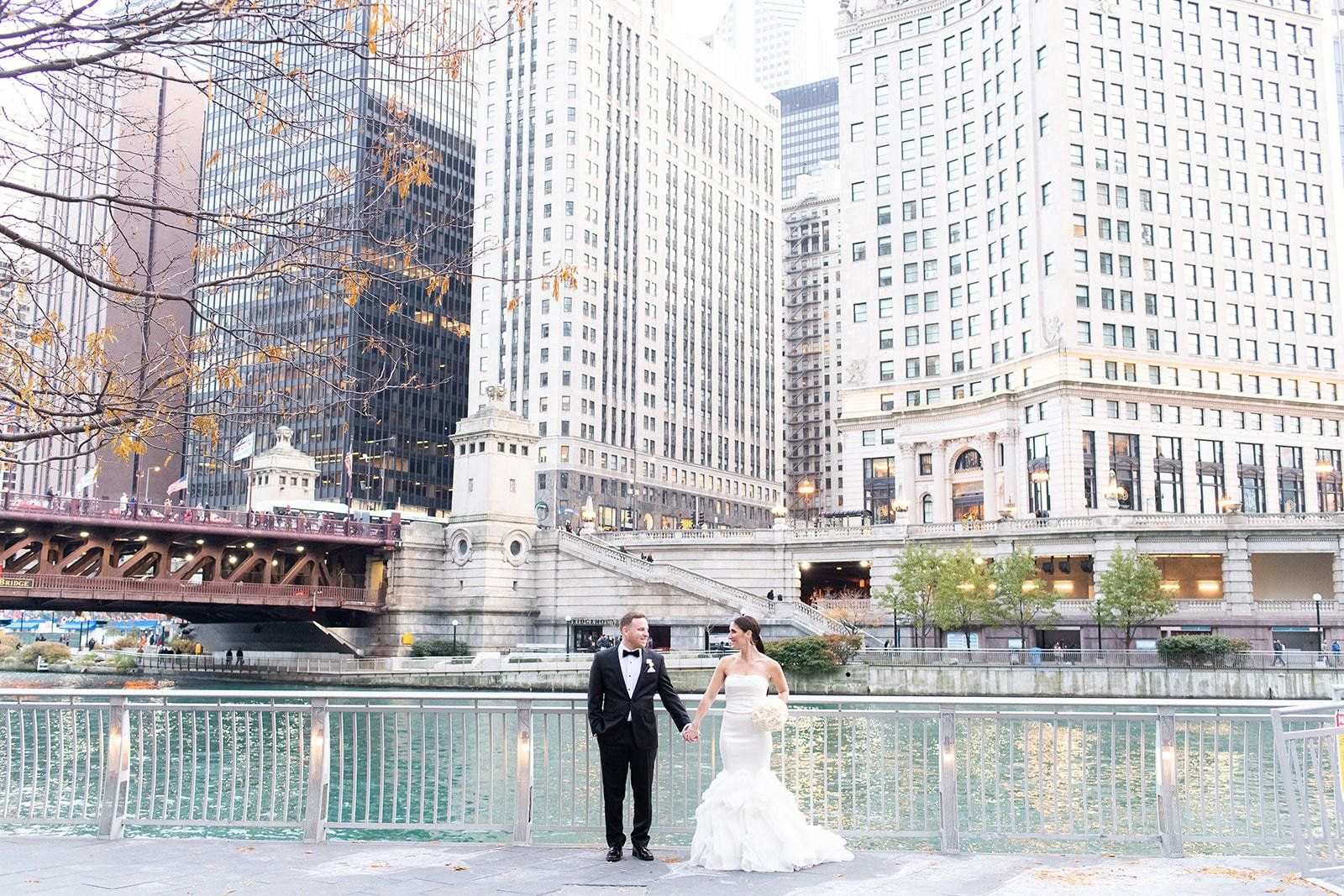 A bride and groom are walking across a bridge in front of a city skyline.