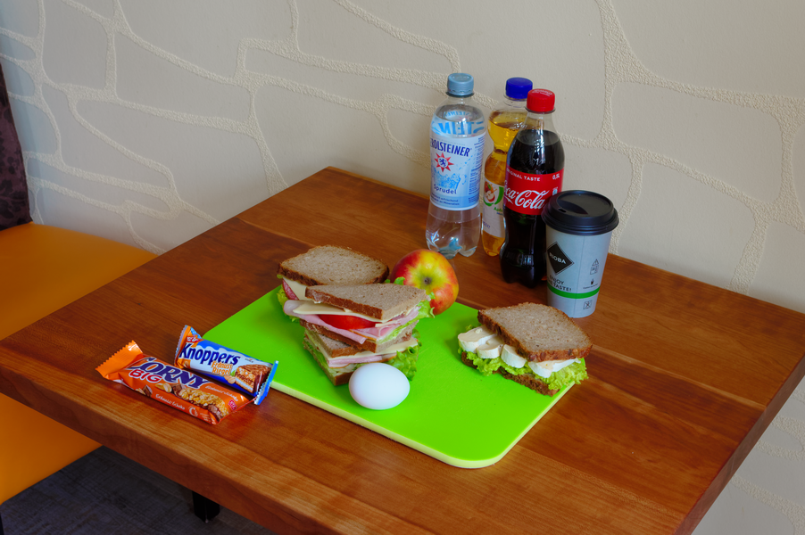 Mittagessen an einem Holztisch: Sandwiches, Getränke (Limonade, Wasser), Apfel, Ei, Schokoriegel.