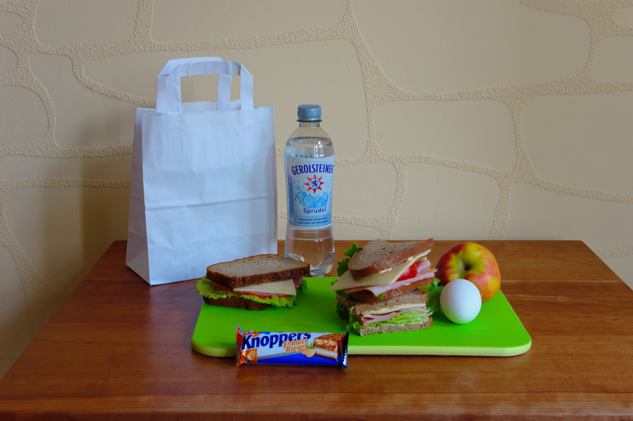 Lunchpaket: weiße Tasche, Wasserflasche, Sandwiches, Apfel, Ei, Müsliriegel auf einem grünen Schneidebrett und ein Holztisch.