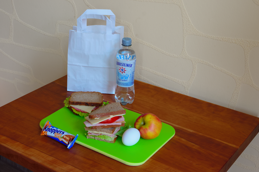 Mittagessen mit Sandwich, Apfel, Ei, Schokoriegel, Wasserflasche und Papiertüte auf einem grünen Schneidebrett.