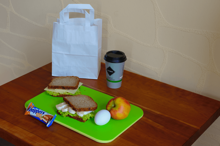 Das Mittagessen auf dem Tisch: Sandwiches, Apfel, hartgekochtes Ei, Energieriegel, Kaffeetasse und eine weiße Papiertüte.