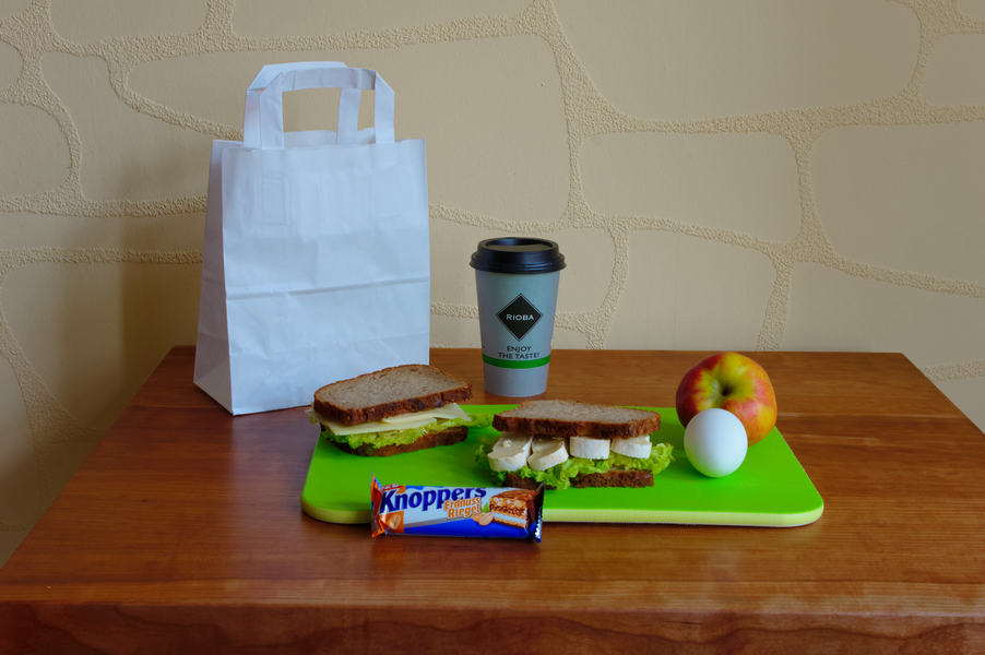 Das Mittagessen auf einem Holztisch umfasst Sandwiches, Kaffee, einen Apfel, ein hartgekochtes Ei und einen Schokoriegel, daneben eine weiße Papiertüte.