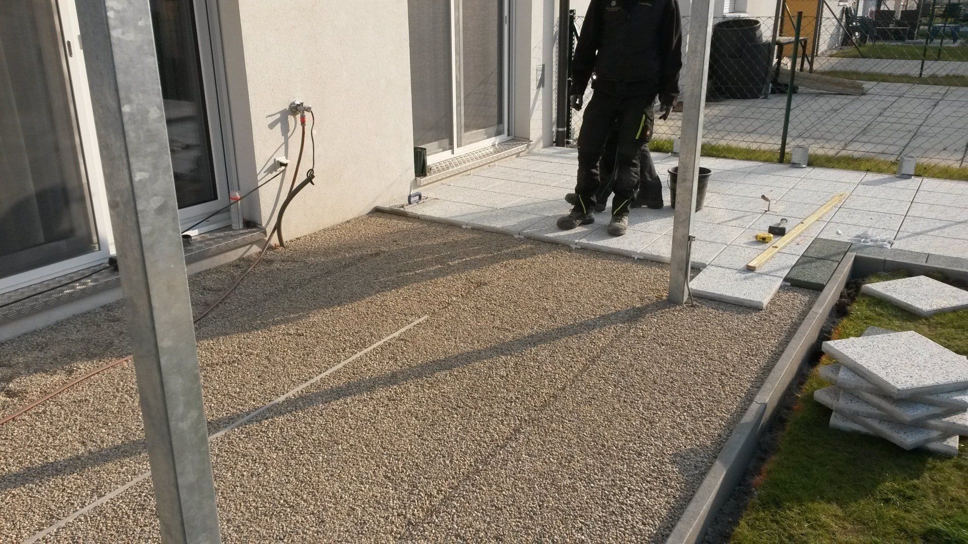 Eine Person in dunkler Kleidung steht auf einer Kieselterrasse neben einem weißen Gebäude und gestapelten Materialien.