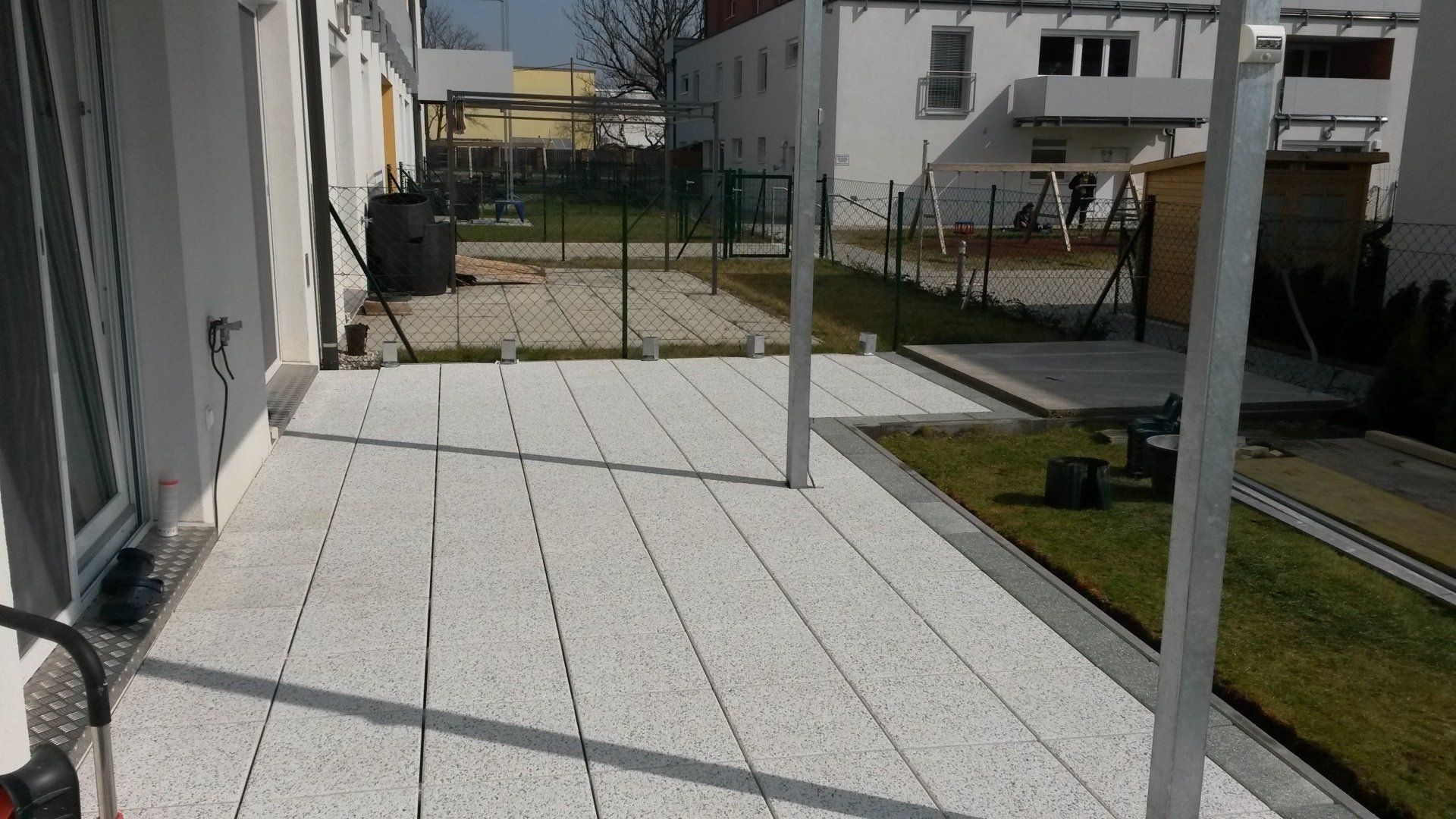 Überdachte Terrasse mit weißem Fliesenboden, Metallpfosten und einem kleinen Rasengarten neben Wohnhäusern.