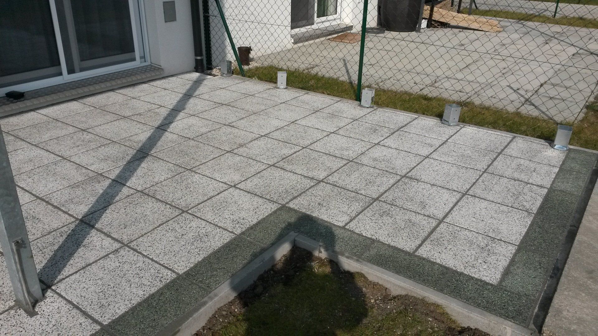 Gepflasterte Terrasse mit quadratischen Fliesen, Maschendrahtzaun und eine kleine Rasenfläche vor dem Haus.