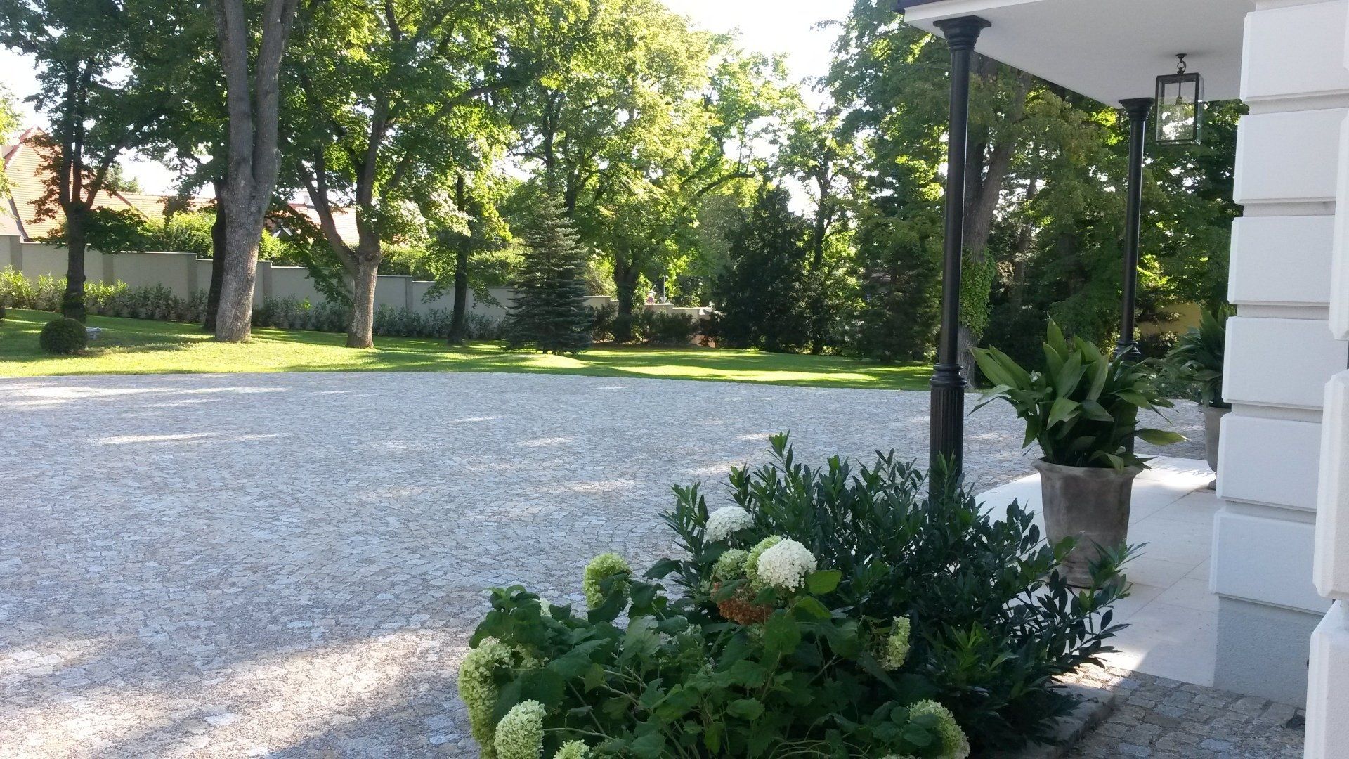 Eine Kiesauffahrt neben einem weißen Haus mit Veranda, Topfpflanzen und sonnenbeschienenen Bäumen im Hintergrund.