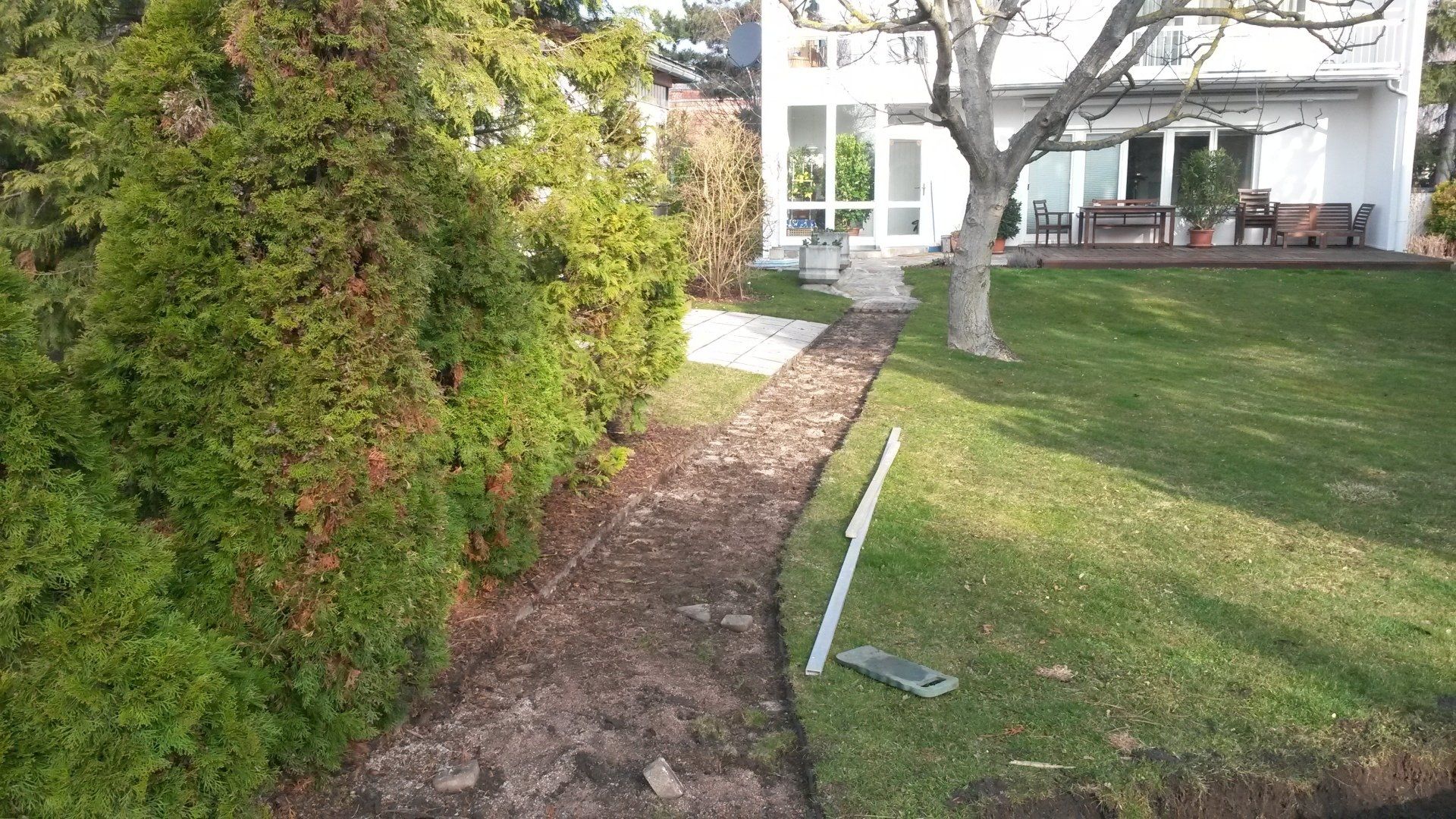 Ein Gartenweg führt neben Hecke und Rasen zu einem weißen Haus mit Terrassenstühlen.