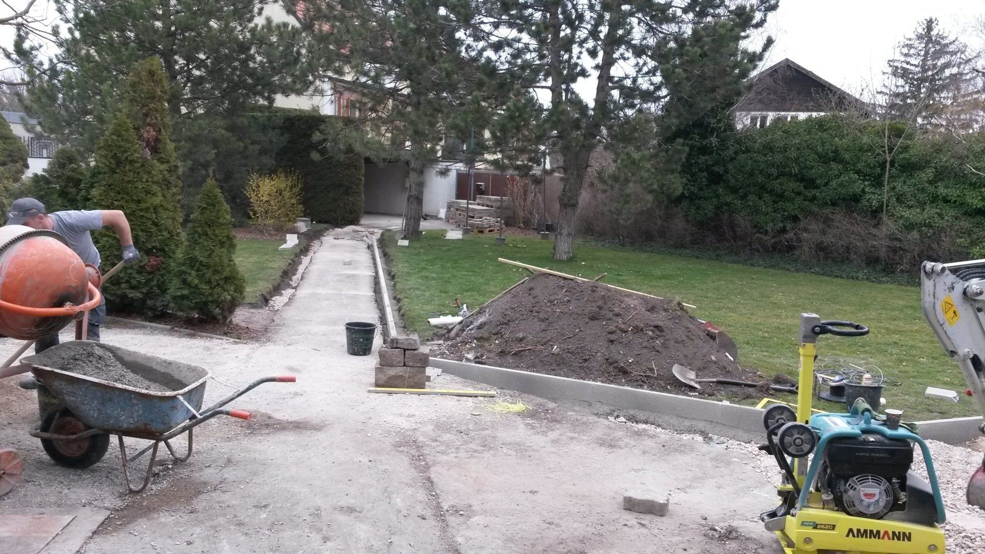 Im Bau befindlicher Gartenweg mit Schubkarre, Kieshaufen und gelber Rüttelplatte auf einem Betonweg.