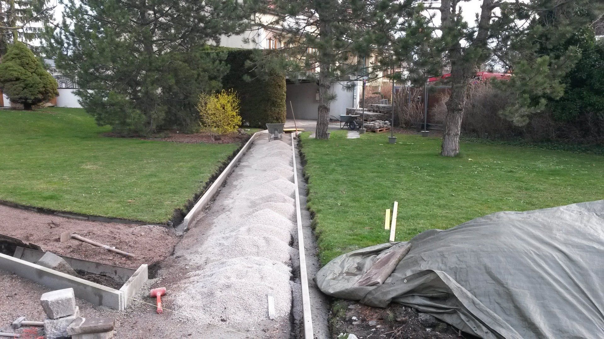Ein schmaler, im Bau befindlicher Betonweg führt durch einen grasbewachsenen Hof zu einem Haus.