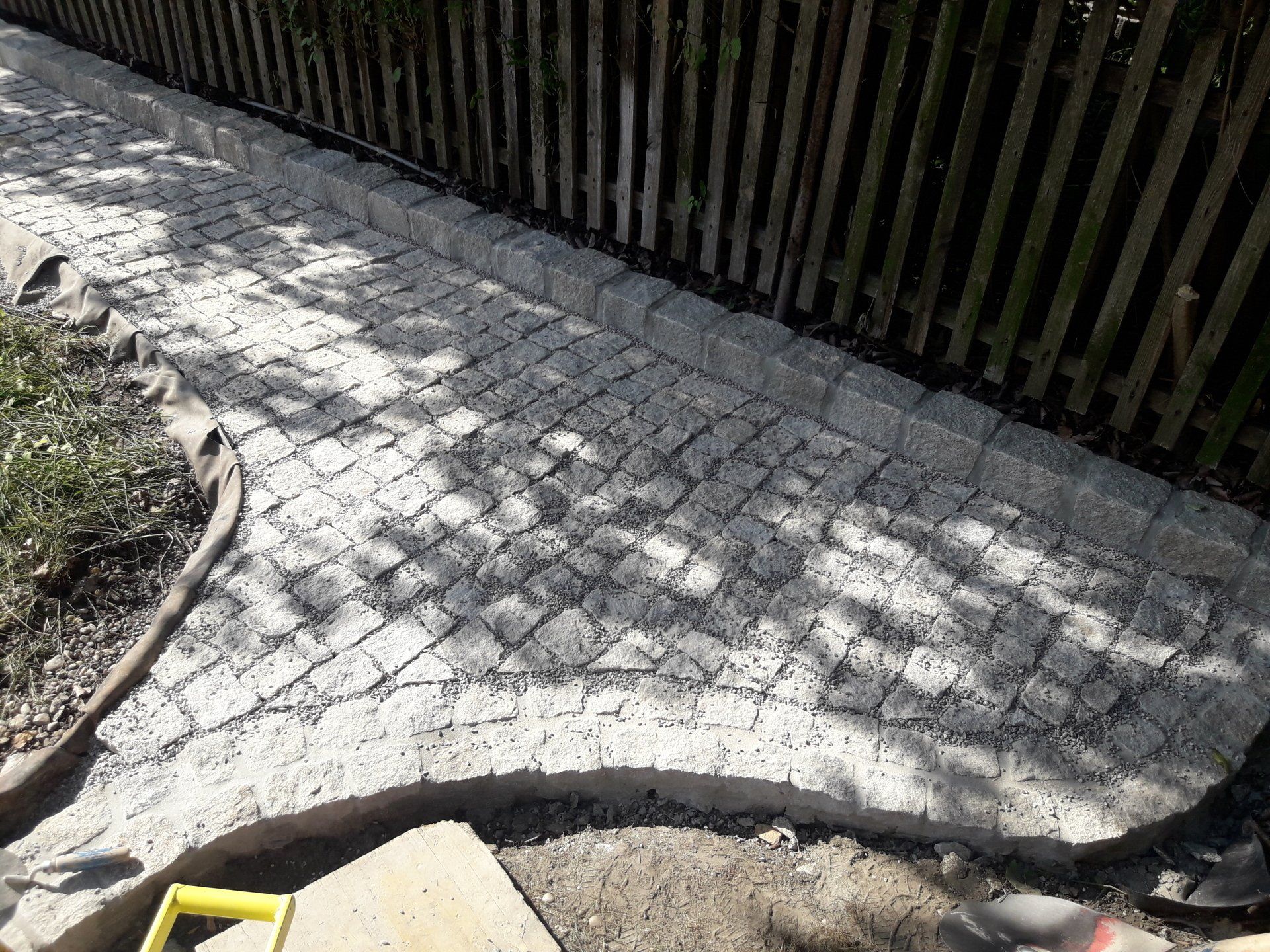 Geschwungener Kopfsteinpflasterweg neben einem dunklen Holzzaun, mit einem gelben Bordstein unten links.