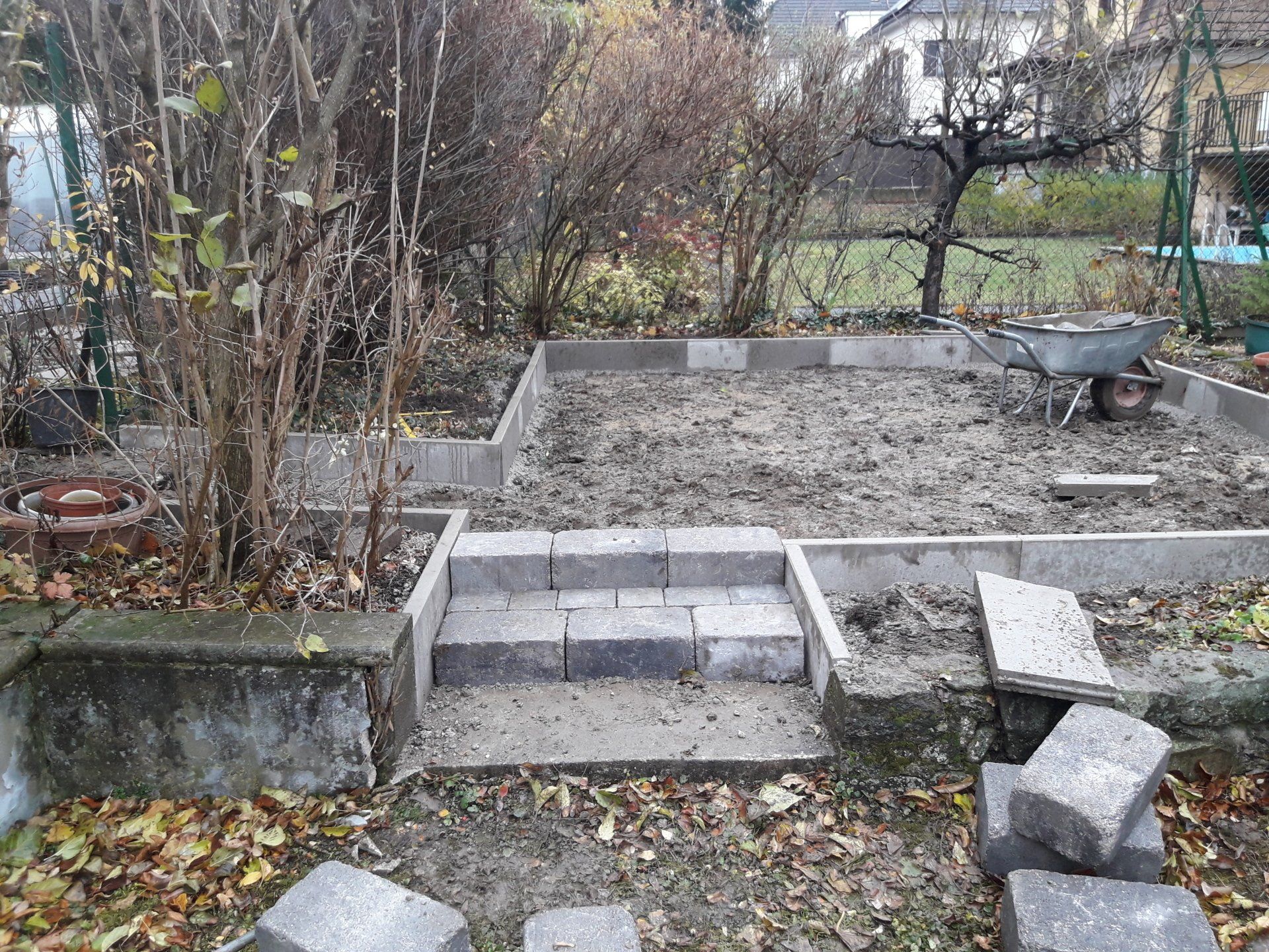 Hinterhofgarten mit erhöhten Betonbeeten, Stufen und einer Schubkarre auf nacktem Boden