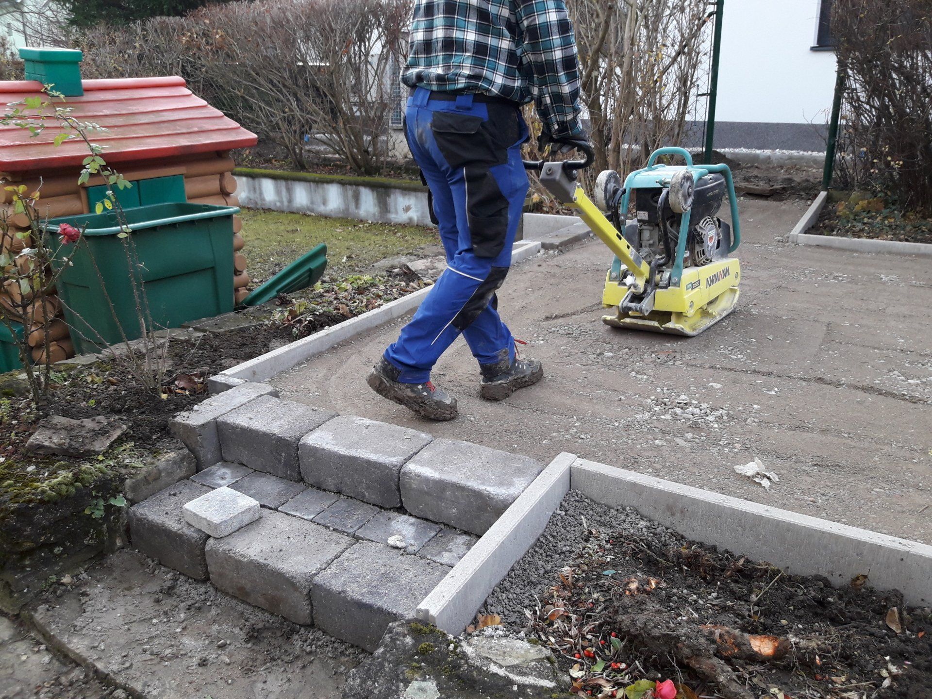 Ein Arbeiter verlegt graue Betonblöcke neben einer Rüttelplatte in einem Hof.