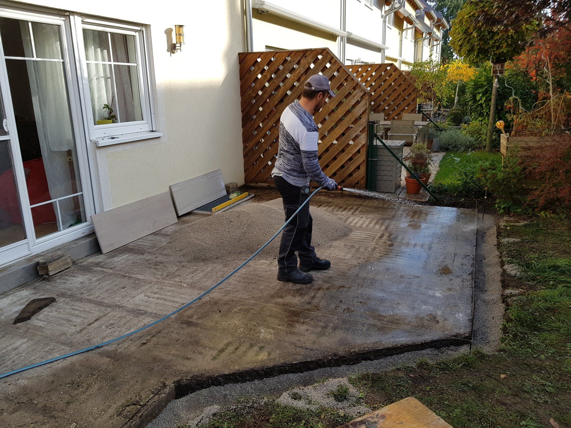 Eine Person reinigt eine Beton-Terrasse neben einem Haus mit einem Hochdruckreiniger; in der Nähe befinden sich ein Garten und ein Zaun.