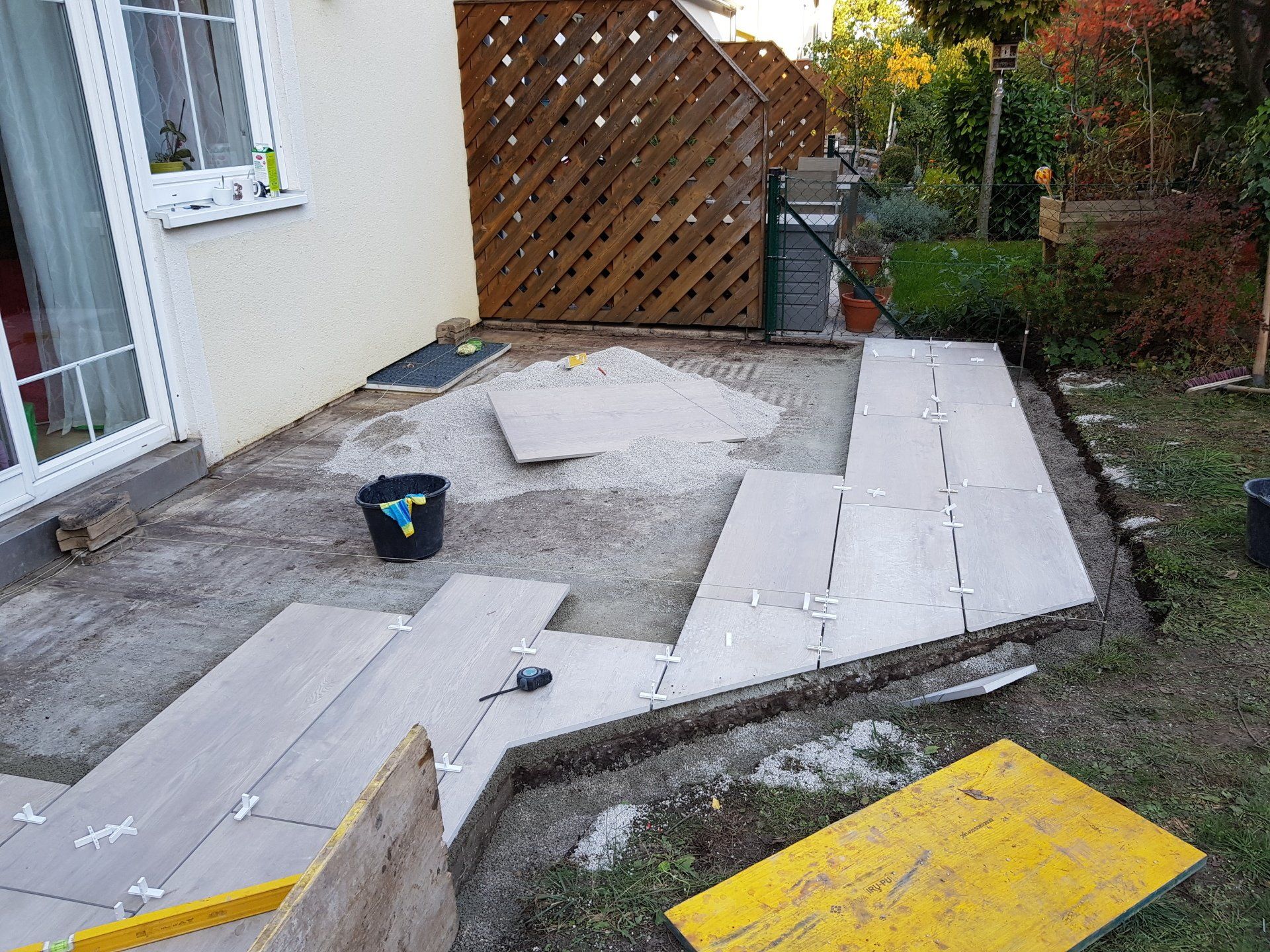 Im Bau befindliche Terrasse im Hinterhof mit Betonplatten, Werkzeugen und einem Holzzaun in der Nähe eines Hauses.