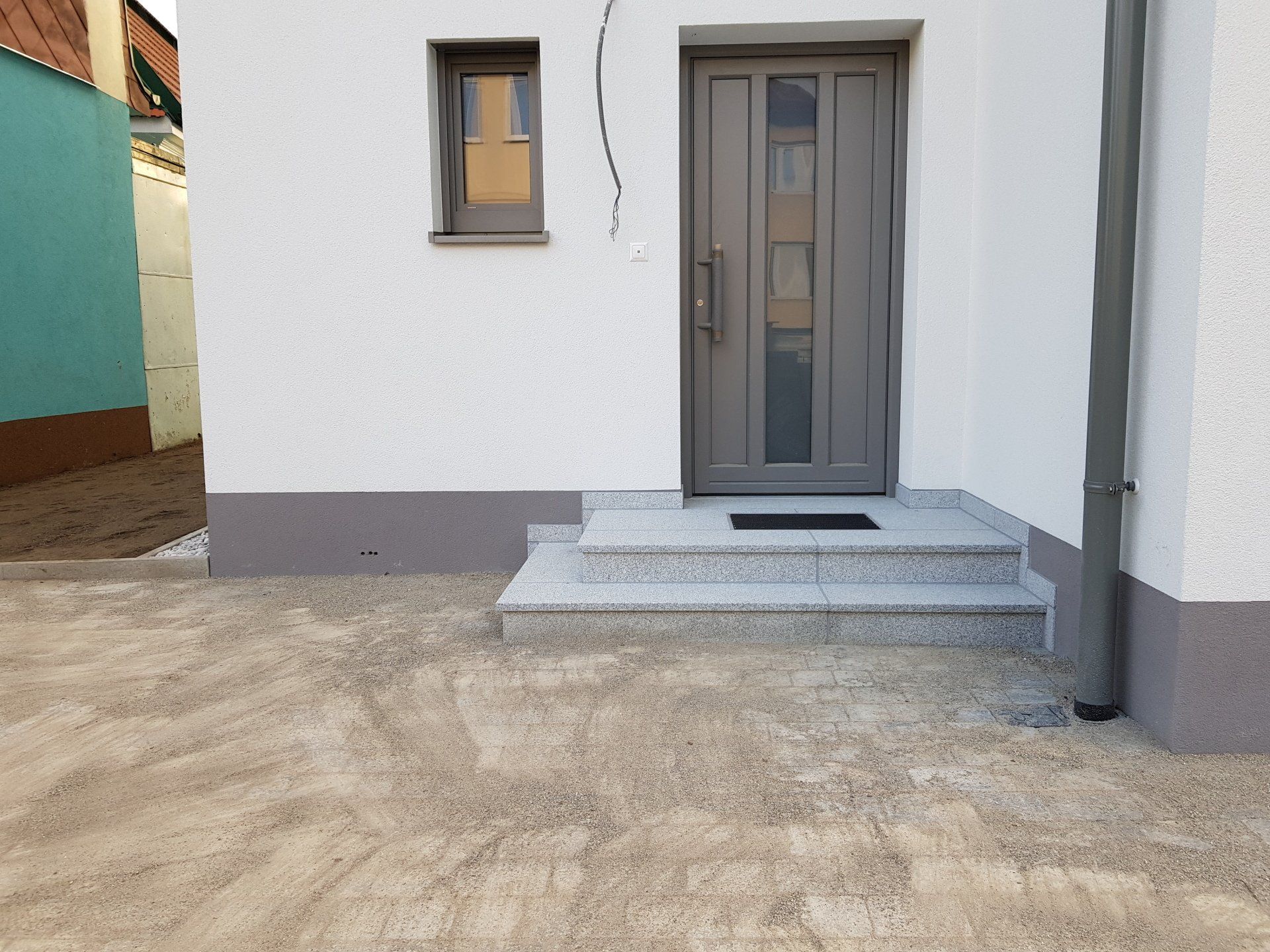 Vordereingang eines weißen Hauses mit grauer Tür, kleinen Stufen und einer betonierten Auffahrt
