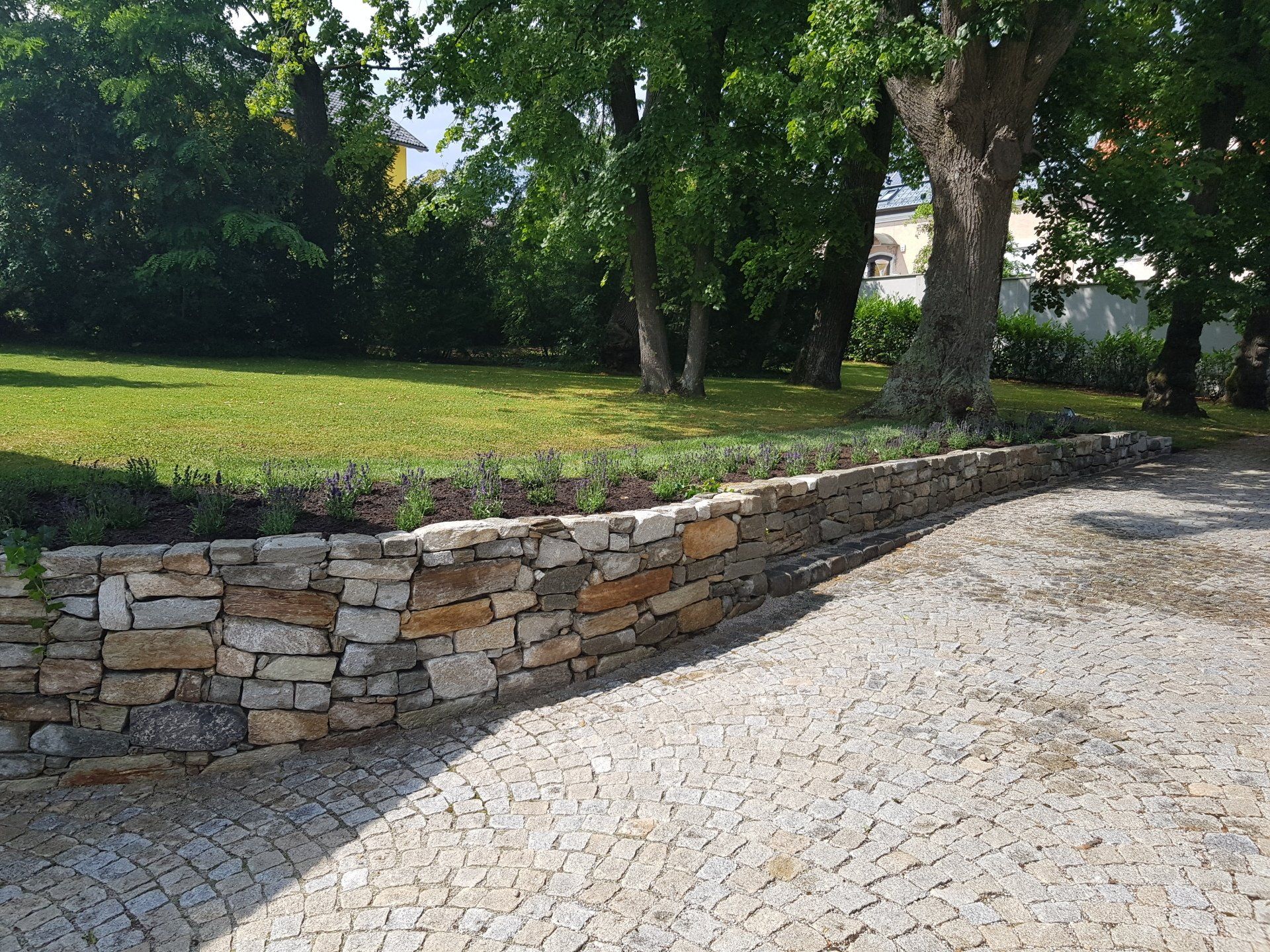 Gebogene Steinmauer neben einem Kiesweg in einem von Bäumen gesäumten Park