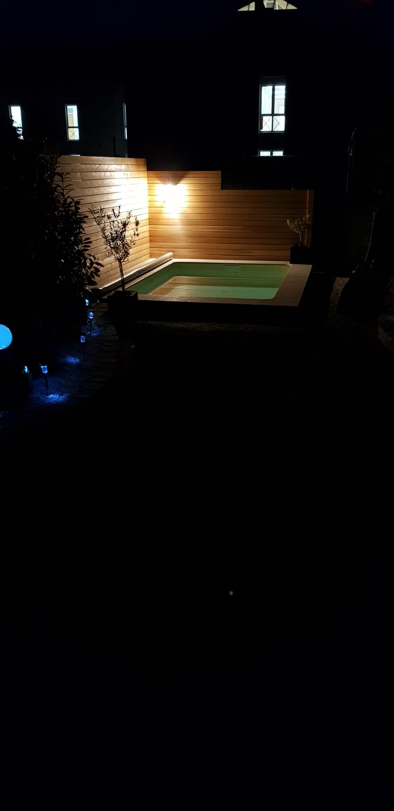Eine schwach beleuchtete Terrasse im Hinterhof mit Holzstufen, ein grün beleuchteter Pool oder Whirlpool und nachts leuchtende Hausfenster.