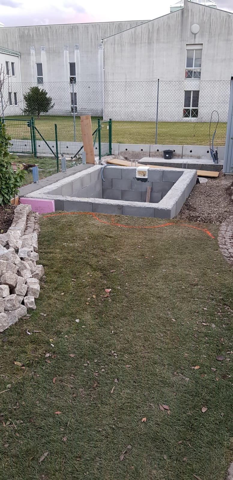 Betonschacht in einem grasbewachsenen Hof, im Hintergrund ein Gebäude und ein Zaun.