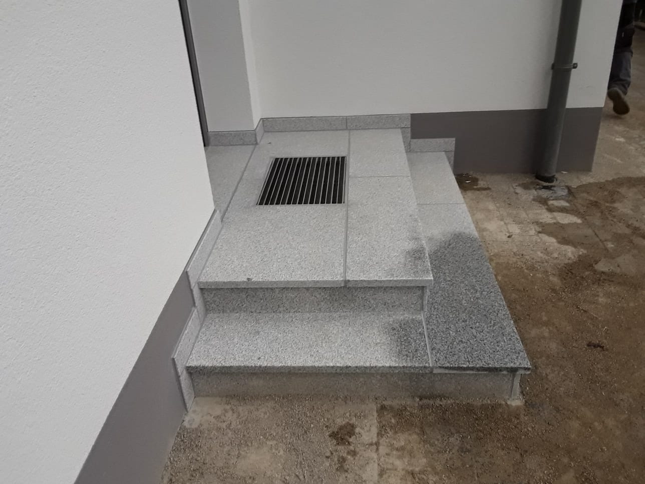 Graue Betonstufen führen zu einem kleinen Metallablaufgitter neben einer weißen Außenwand.