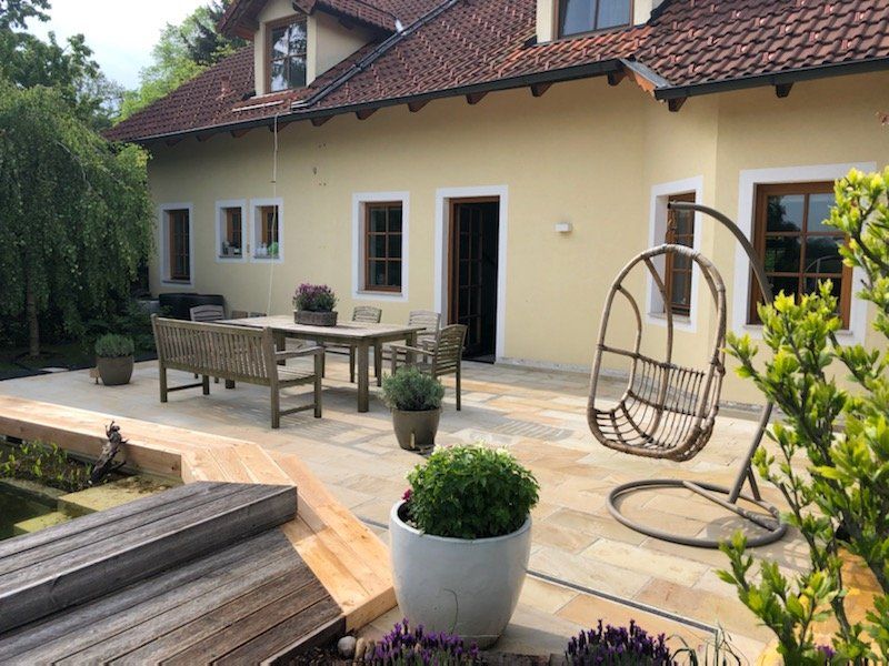 Terrasse mit Korbmöbeln, Topfpflanzen und modernen Stühlen neben einem cremefarbenen Haus mit rotem Dach