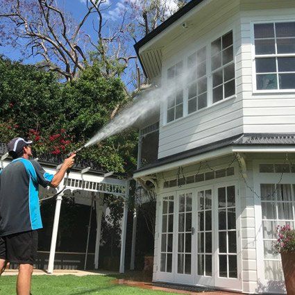 Person pressure-washing a two-story white house beside a garden and carport