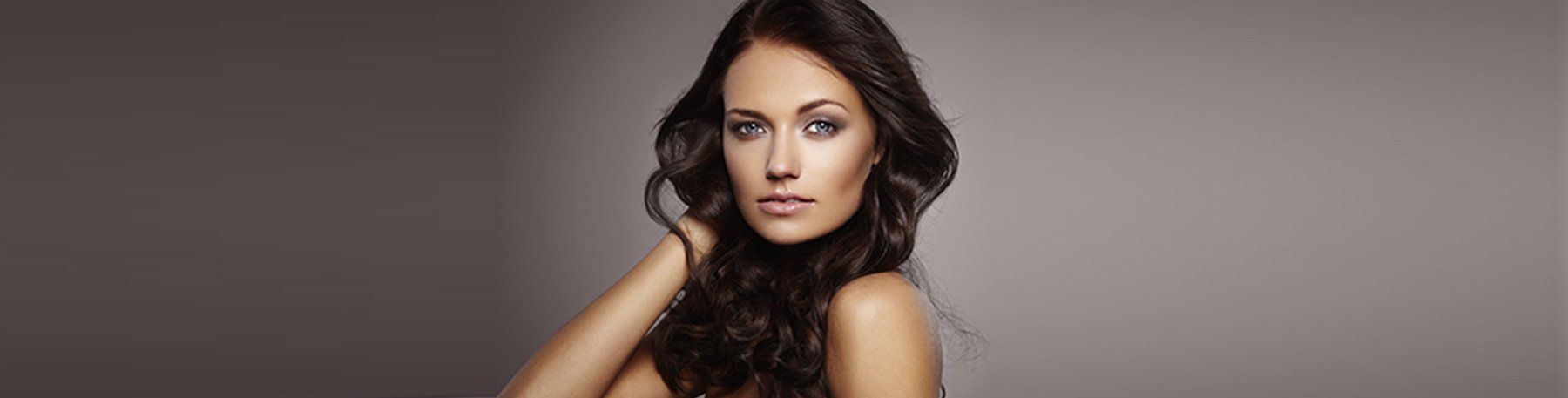 A woman with long dark wavy hair, hand near her face, looking forward, against a gray background.
