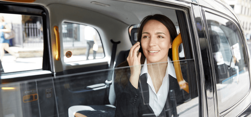 A lady in a black jacket and white shirt, sitting in the back of a taxi, talking on a mobile phone