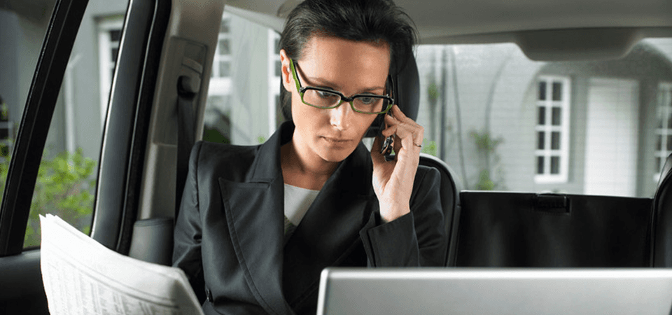 A lady sitting in a car holding a newspaper, looking at a laptop screen, a phone to her ear