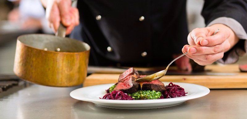 Un chef dresse un plat gastronomique avec sa sauce dans une cuisine ; gros plan sur ses mains et une assiette blanche.