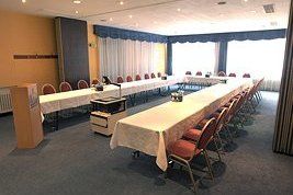 Sala de conferencias con mesas en forma de U, sillas rojas y grandes ventanales con cortinas azules.