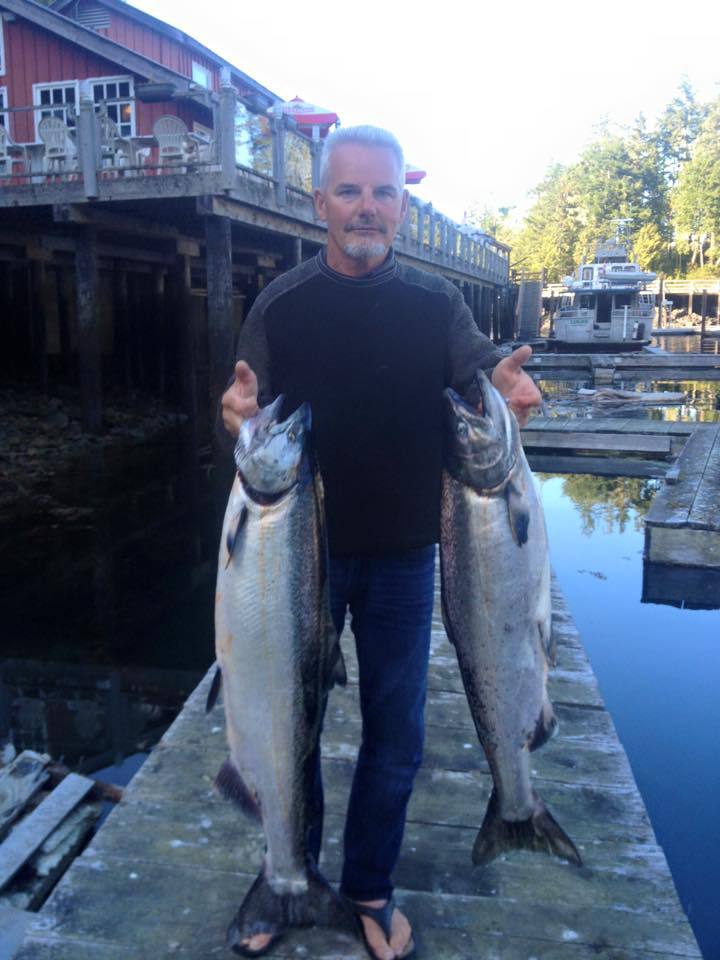 Man standing on a dock holding two large silver fish, with a marina and boats in the background