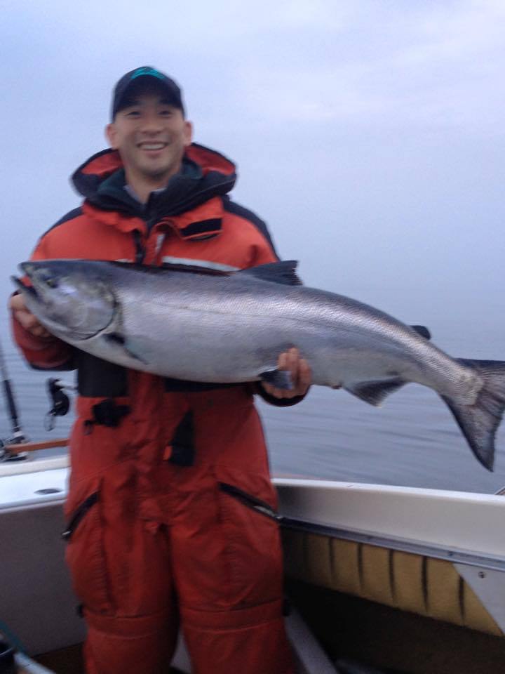Person in red fishing gear holds a large silver fish on a boat in calm water.