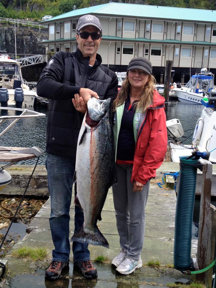 Two anglers posing at a marina, holding a large silver fish in front of docked boats.