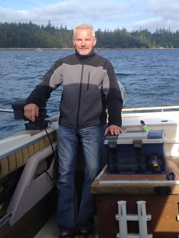 Man standing on a boat deck by the water, wearing a gray jacket and jeans, with forested shoreline behind him