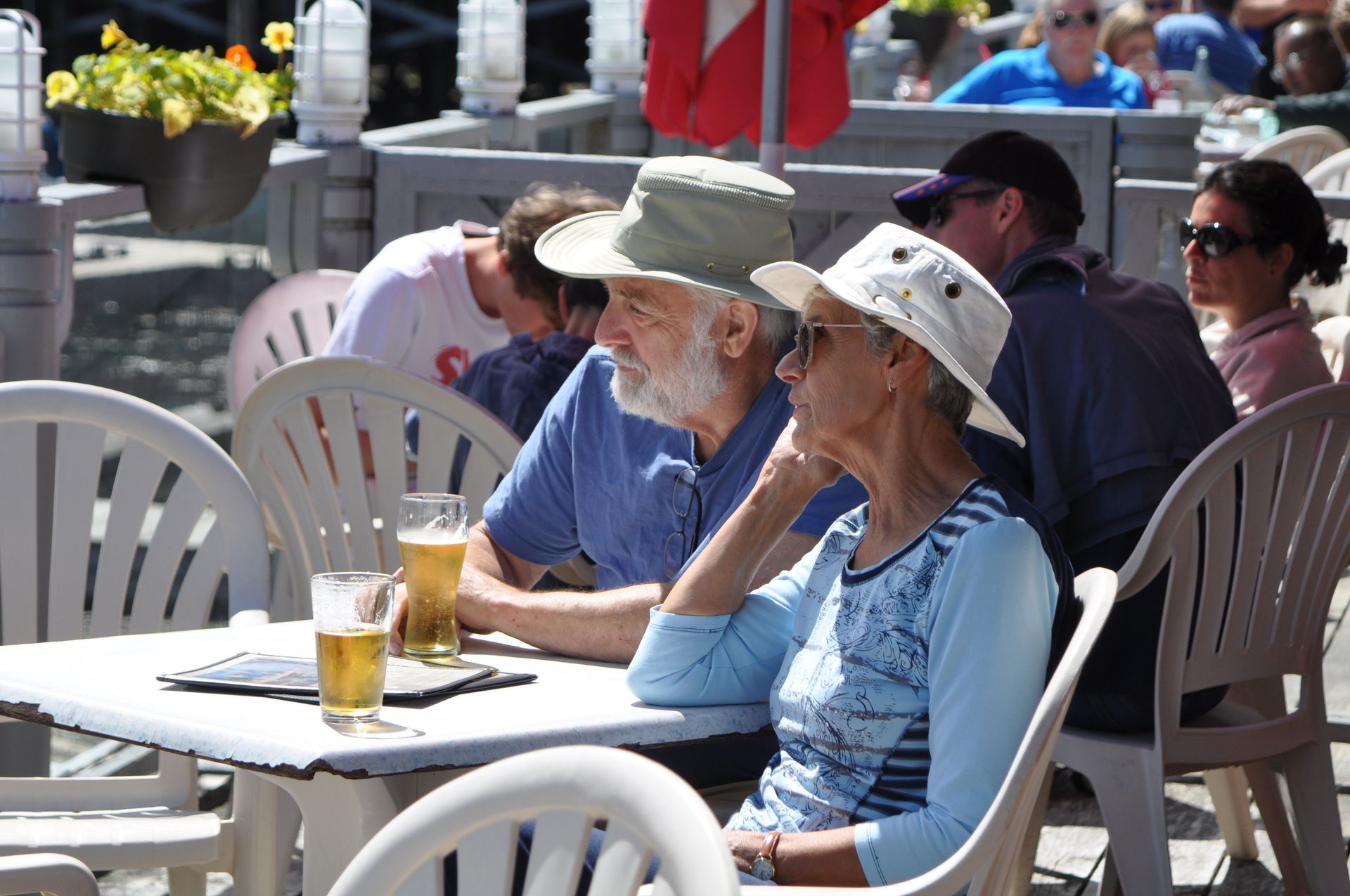 People sitting at outdoor café tables in sunlight, with drinks and hats on a patio.