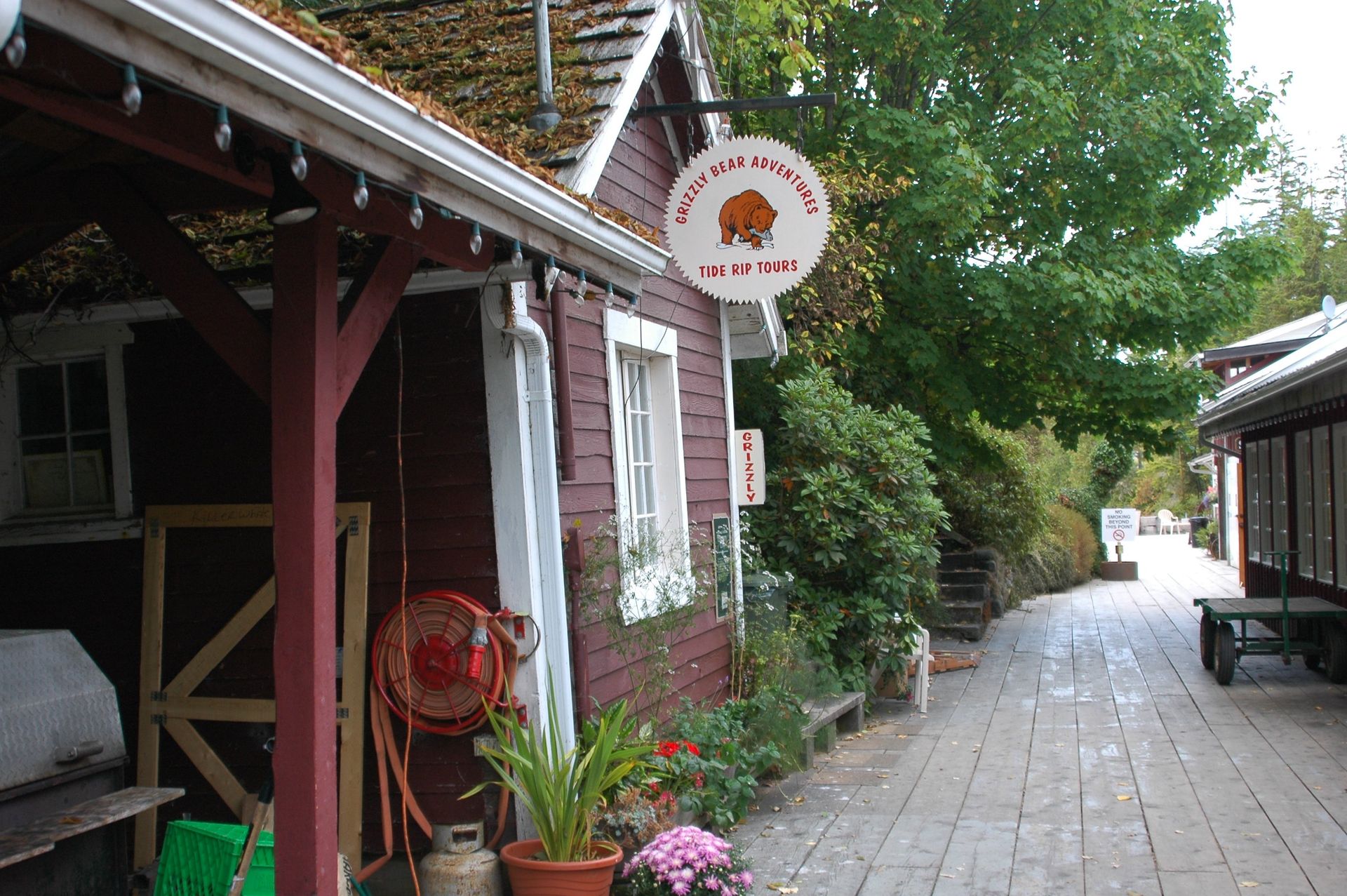 Narrow wooden walkway between rustic red buildings with potted plants and a hanging sign.