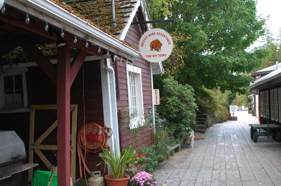 Narrow wooden walkway between rustic shops, with potted plants and a round hanging sign.